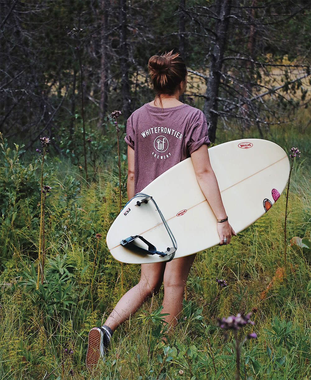 Femme dans une foret portant un t-shirt whitefrontier avec une planche de surf
